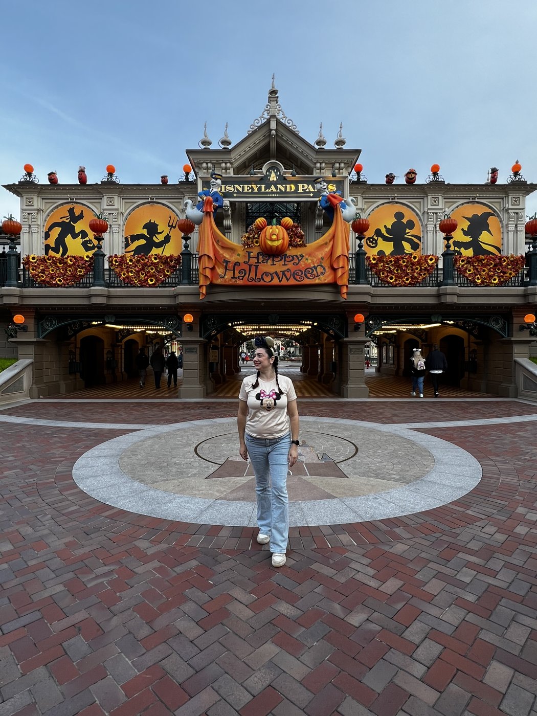 Gabriela Bordin na entrada da Disneyland Paris decorada para o Halloween, com abóboras e o letreiro Happy Halloween